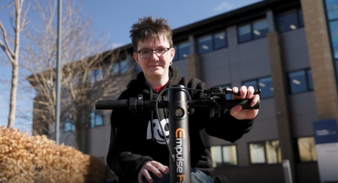 Chelsea Douglas outdoors on a sunny day, smiling while holding the handlebars of her powered wheelchair attachment, with a modern building and trees in the background.