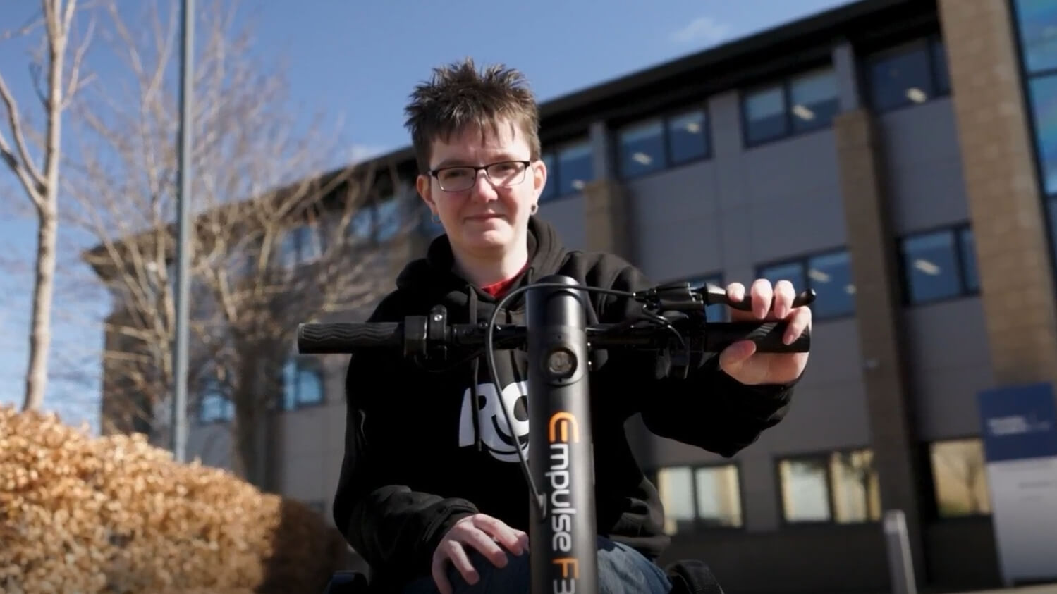 Chelsea Douglas outdoors on a sunny day, smiling while holding the handlebars of her powered wheelchair attachment, with a modern building and trees in the background.