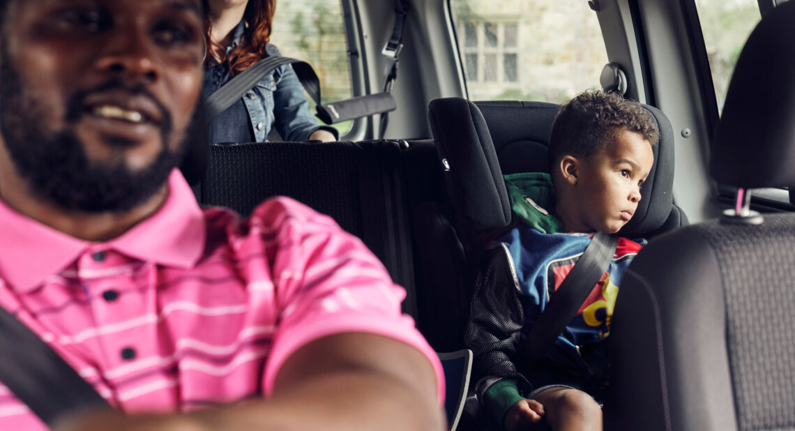 A man drives a car with a woman in the back seat and a young child sitting safely in a car seat, secured with a seatbelt and looking out the window.
