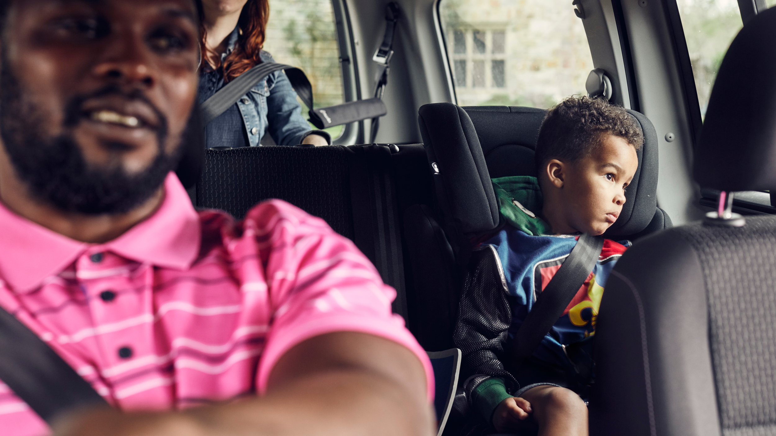 A man drives a car with a woman in the back seat and a young child sitting safely in a car seat, secured with a seatbelt and looking out the window.