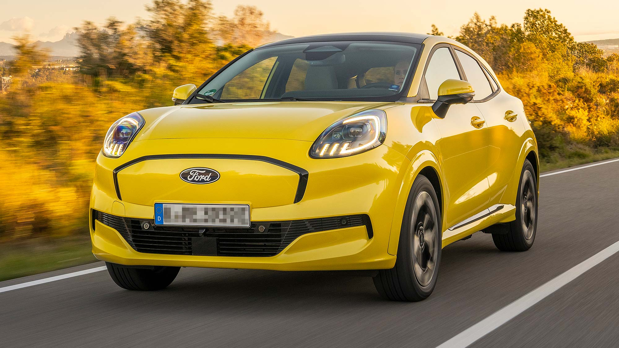Bright yellow Ford Puma Gen-E electric crossover driving on a scenic road at sunset, with golden fields in the background.