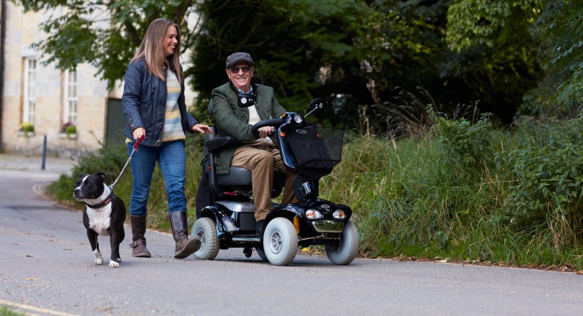 A man uses a mobility scooter along a quiet path beside greenery, accompanied by a woman walking beside him holding a dog on a lead. They appear to be enjoying a relaxed conversation outdoors.