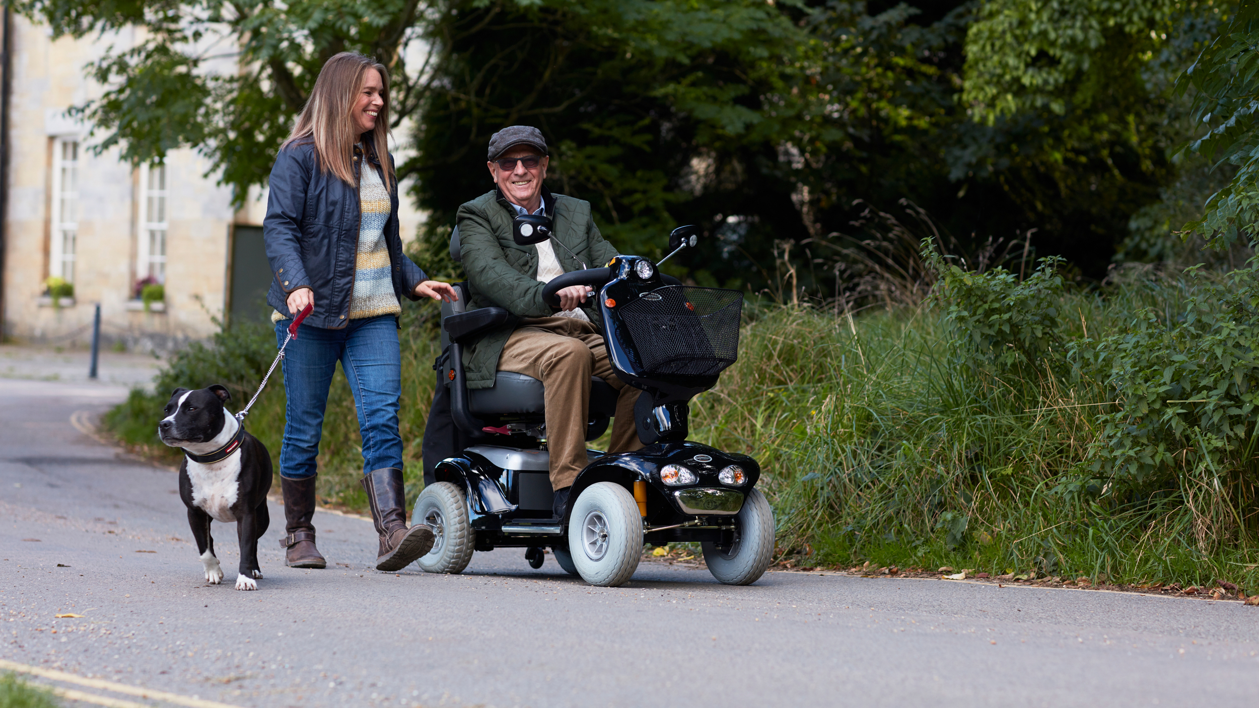 A man uses a mobility scooter along a quiet path beside greenery, accompanied by a woman walking beside him holding a dog on a lead. They appear to be enjoying a relaxed conversation outdoors.
