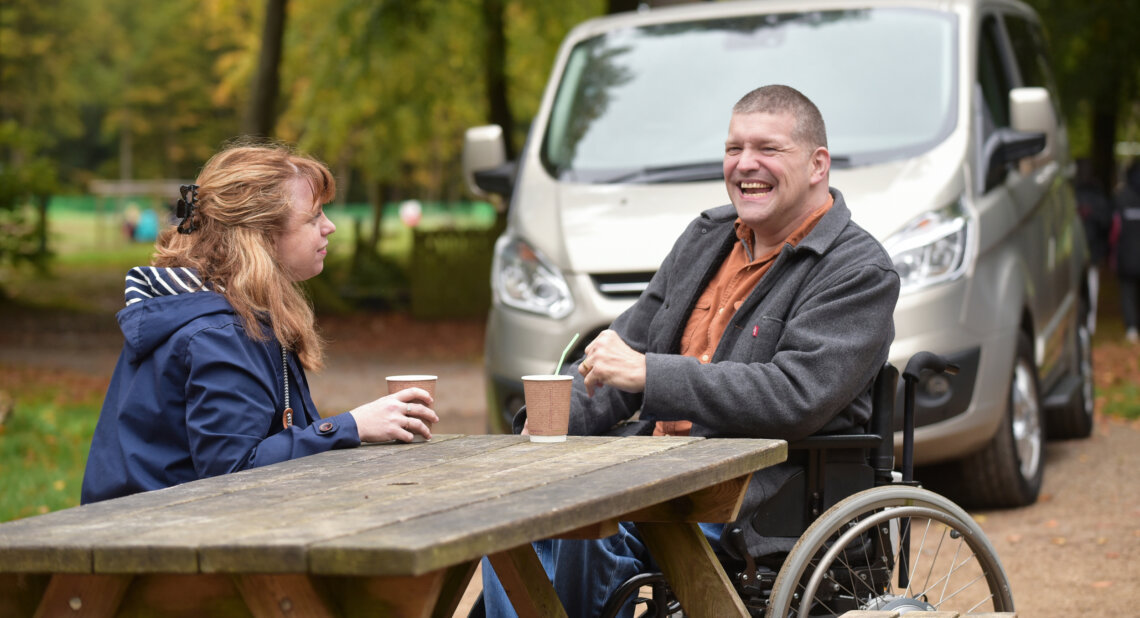 A man and woman laugh on a park bench with Wheelchair Accessible Vehicle (WAV) in the background