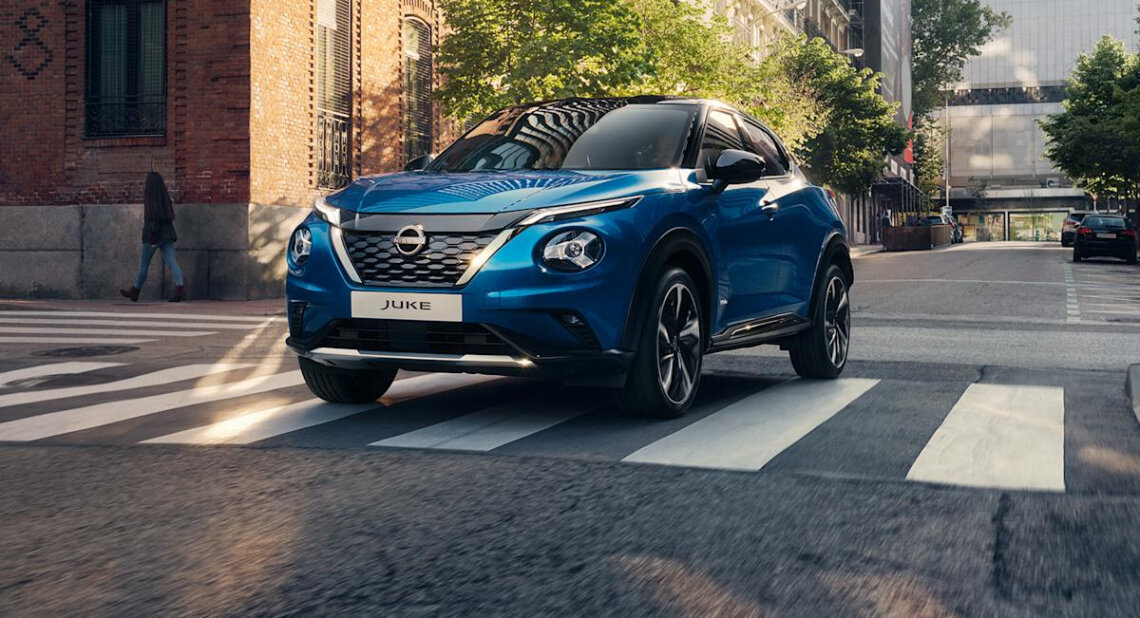 A blue Nissan Juke driving through a city street, crossing a pedestrian crossing with buildings and trees in the background.