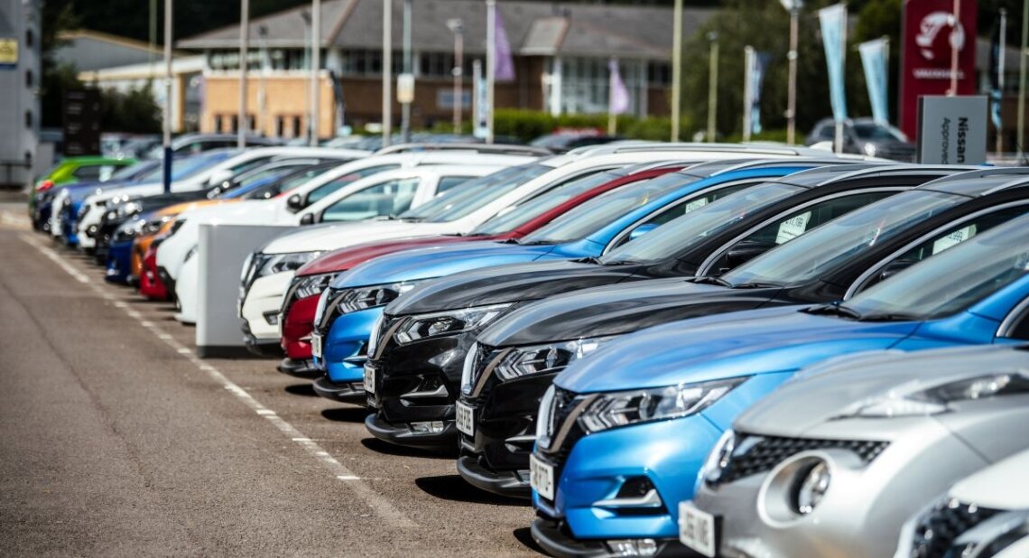 A row of cars parked outside a dealership. The vehicles are lined up neatly, with the dealership sign just visible.