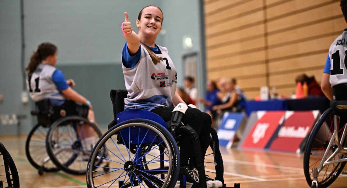A young wheelchair basketball player wearing a blue and white jersey smiles and gives a thumbs up during a game, with teammates visible behind on the court.