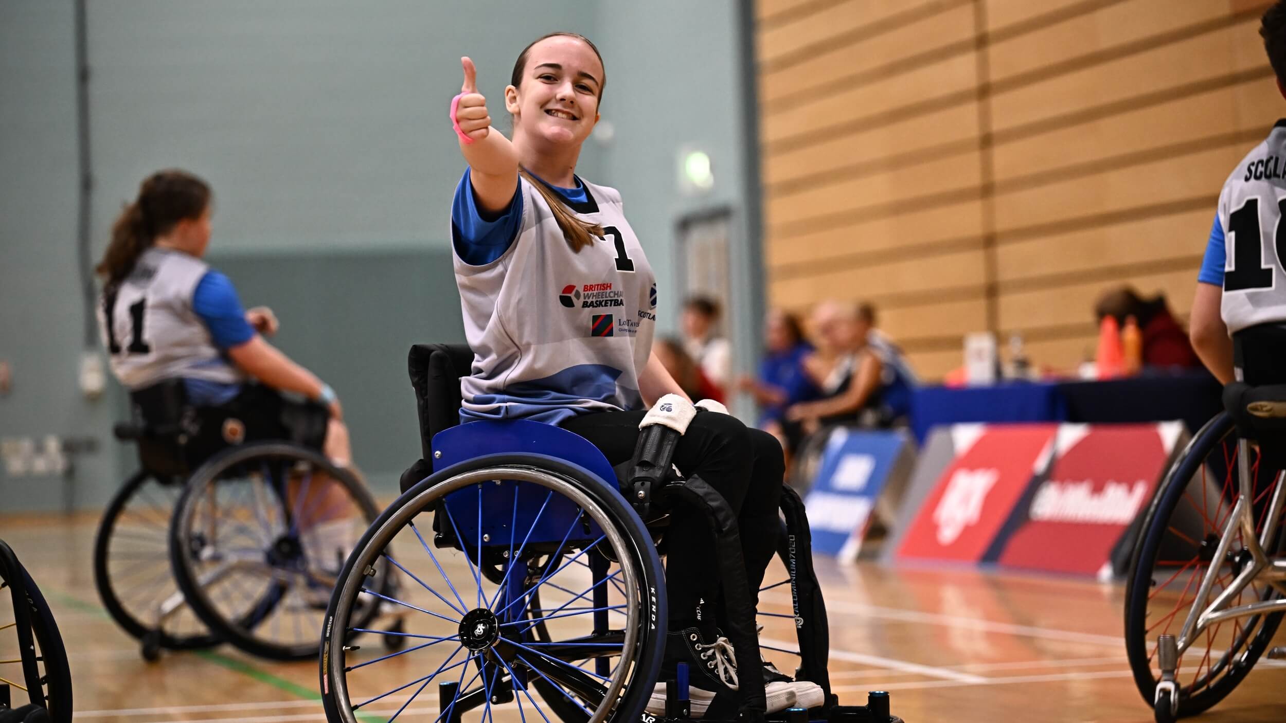 A young wheelchair basketball player wearing a blue and white jersey smiles and gives a thumbs up during a game, with teammates visible behind on the court.