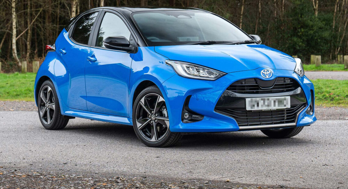 A blue Toyota Yaris hybrid hatchback with black roof is parked on a tarmac surface near a wooded area.