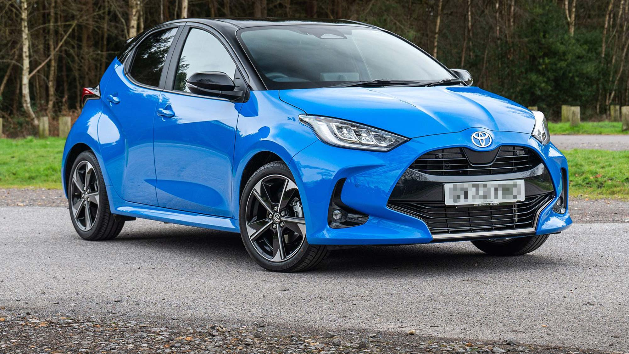 A blue Toyota Yaris hybrid hatchback with black roof is parked on a tarmac surface near a wooded area.