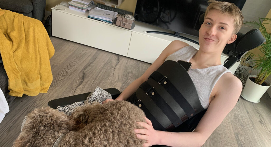 A young person sitting in a wheelchair at home, smiling while resting a hand on a curly-haired dog on their lap. They are wearing a light grey sleeveless top and a black chest support brace, with a television and plant visible in the background.