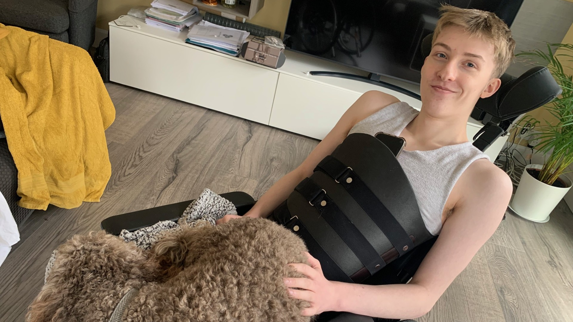 A young person sitting in a wheelchair at home, smiling while resting a hand on a curly-haired dog on their lap. They are wearing a light grey sleeveless top and a black chest support brace, with a television and plant visible in the background.