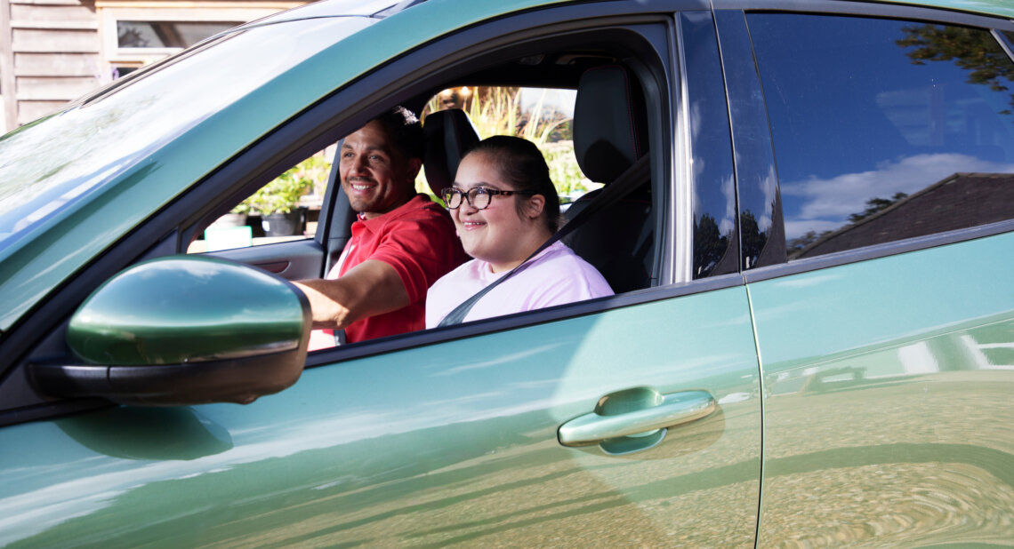 A man and a young girl sit smiling in the front seats of a green car parked outdoors. The man in a red shirt is in the driver’s seat, while the girl beside him wears glasses and a pale pink top. Sunlight reflects off the car’s shiny exterior, and a wooden building is visible in the background.