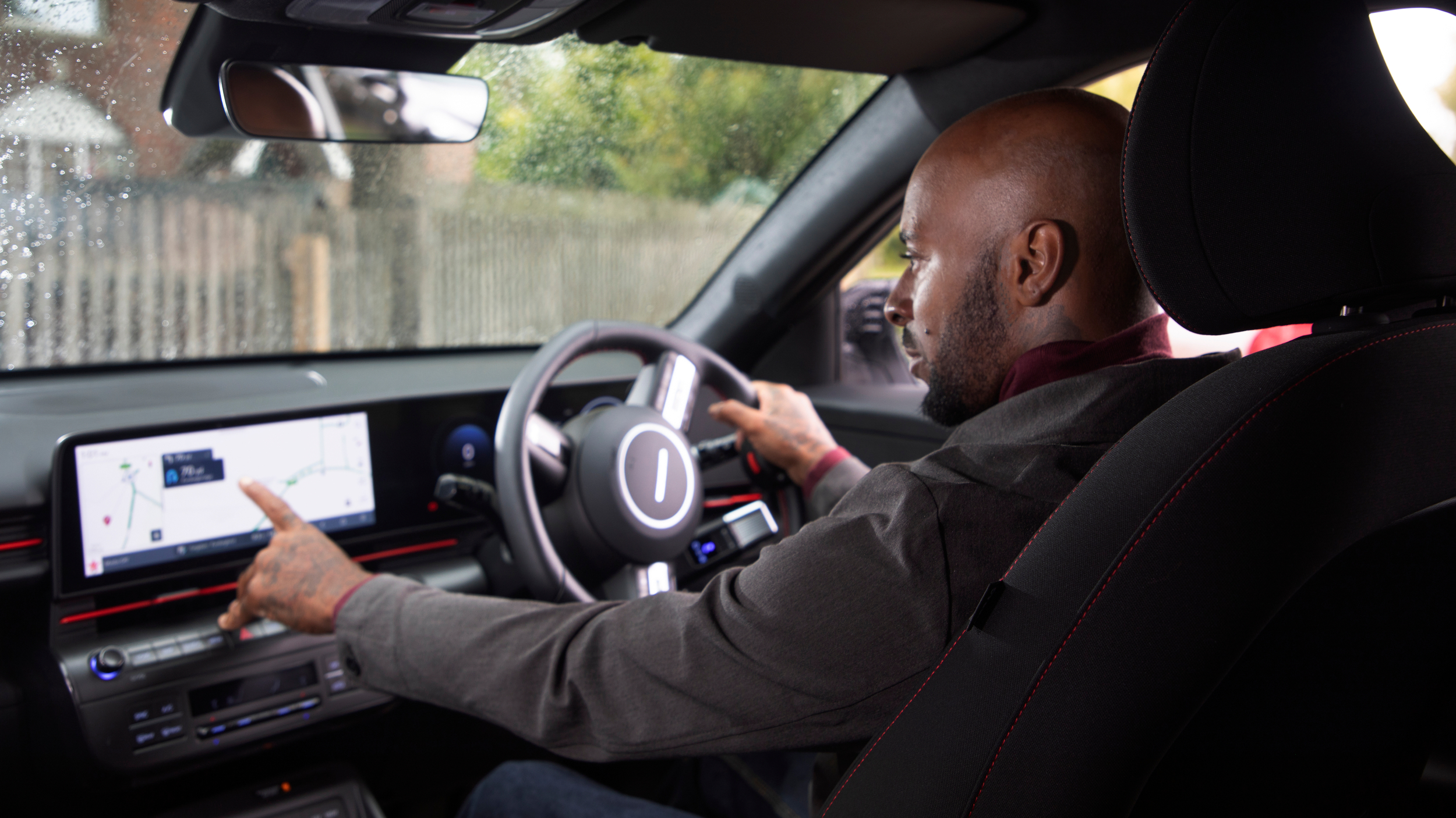 A man seated in the driver’s seat of a car uses the touchscreen navigation system on the dashboard. This is more commonly known as the car infotainment system. It is daytime, and the car windows show raindrops from wet weather outside.