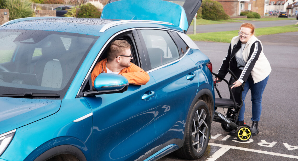 A man in an orange jacket sits in the driver’s seat of a blue car with the boot open, smiling toward a woman who is loading a folded wheelchair into the back. The scene takes place in a car park on a cloudy day, with houses and greenery in the background.