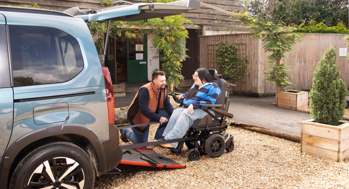 A man kneels beside a woman in a powered wheelchair at the rear of a wheelchair accessible vehicle, with the ramp lowered onto a gravel driveway. They are smiling at each other outside a building surrounded by plants and wooden fencing.