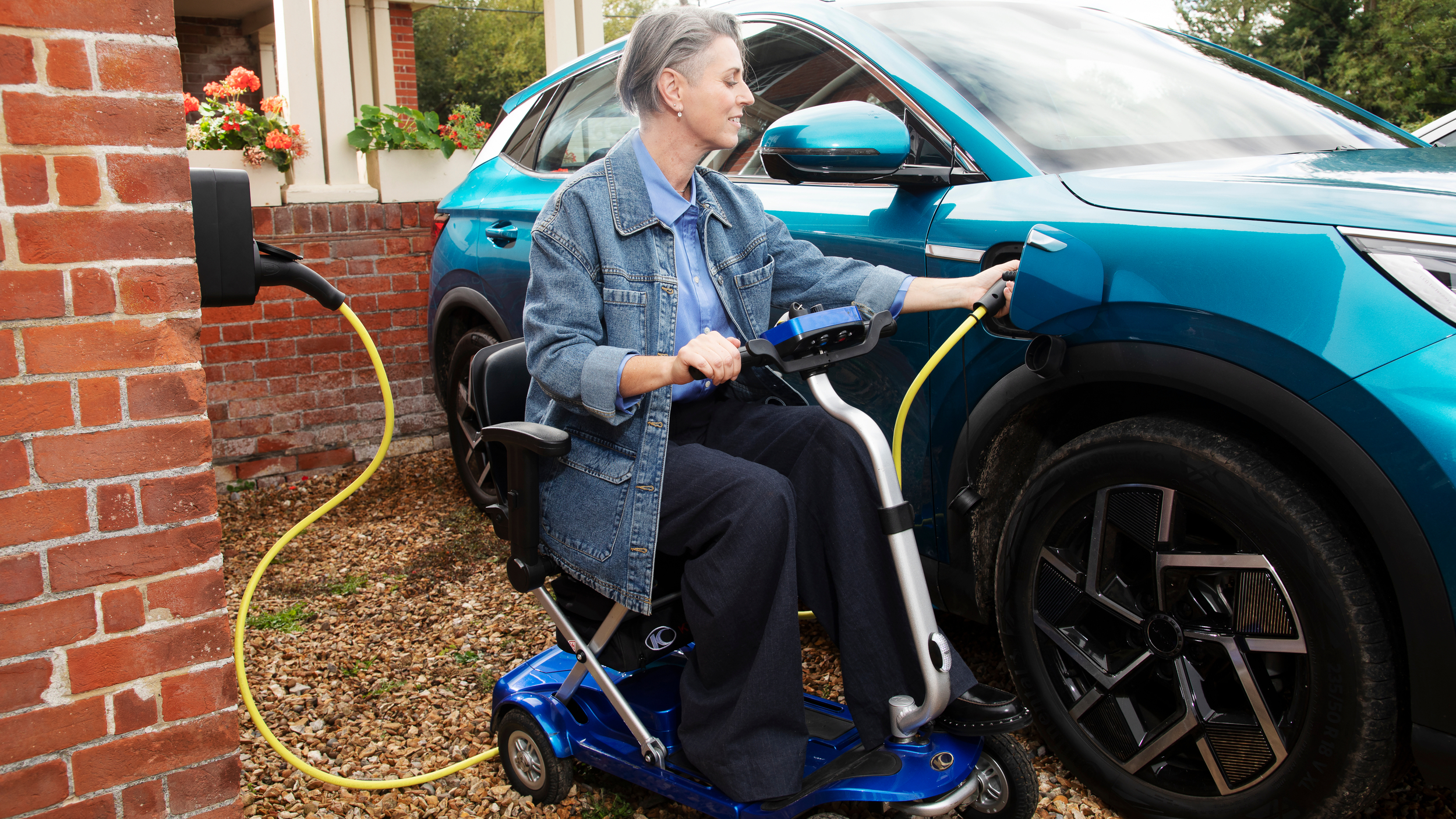 Woman sitting on a blue mobility scooter outside a brick house, plugging a yellow charging cable into a blue electric car parked on the driveway.