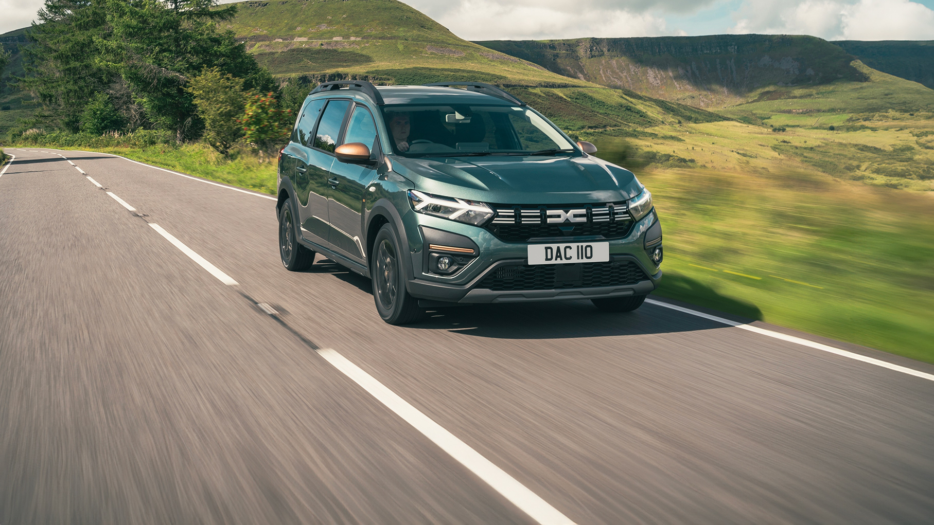 A green Dacia Jogger driving on a road with a view of cliffs in the distance, under a bright sky.