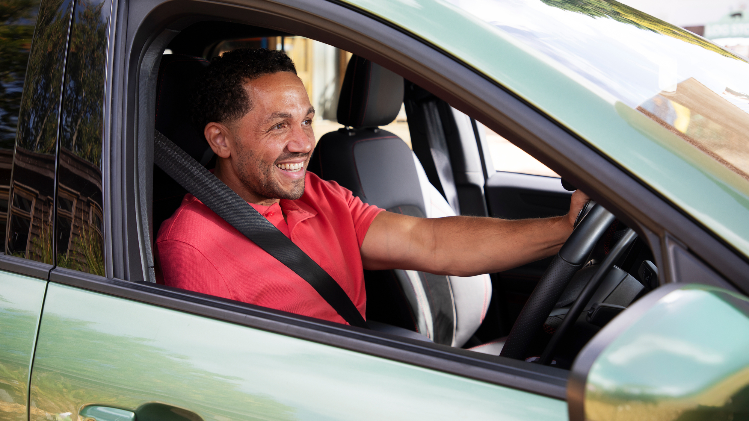 A man smiling while sitting in the driver’s seat of a green car, wearing a red polo shirt and a seatbelt, with sunlight reflecting off the car’s window.
