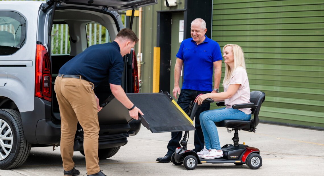 A Wheelchair Accessible Vehicle (WAV) expert lowers a ramp at the rear of the vehicle for a lady in a wheelchair and a man.