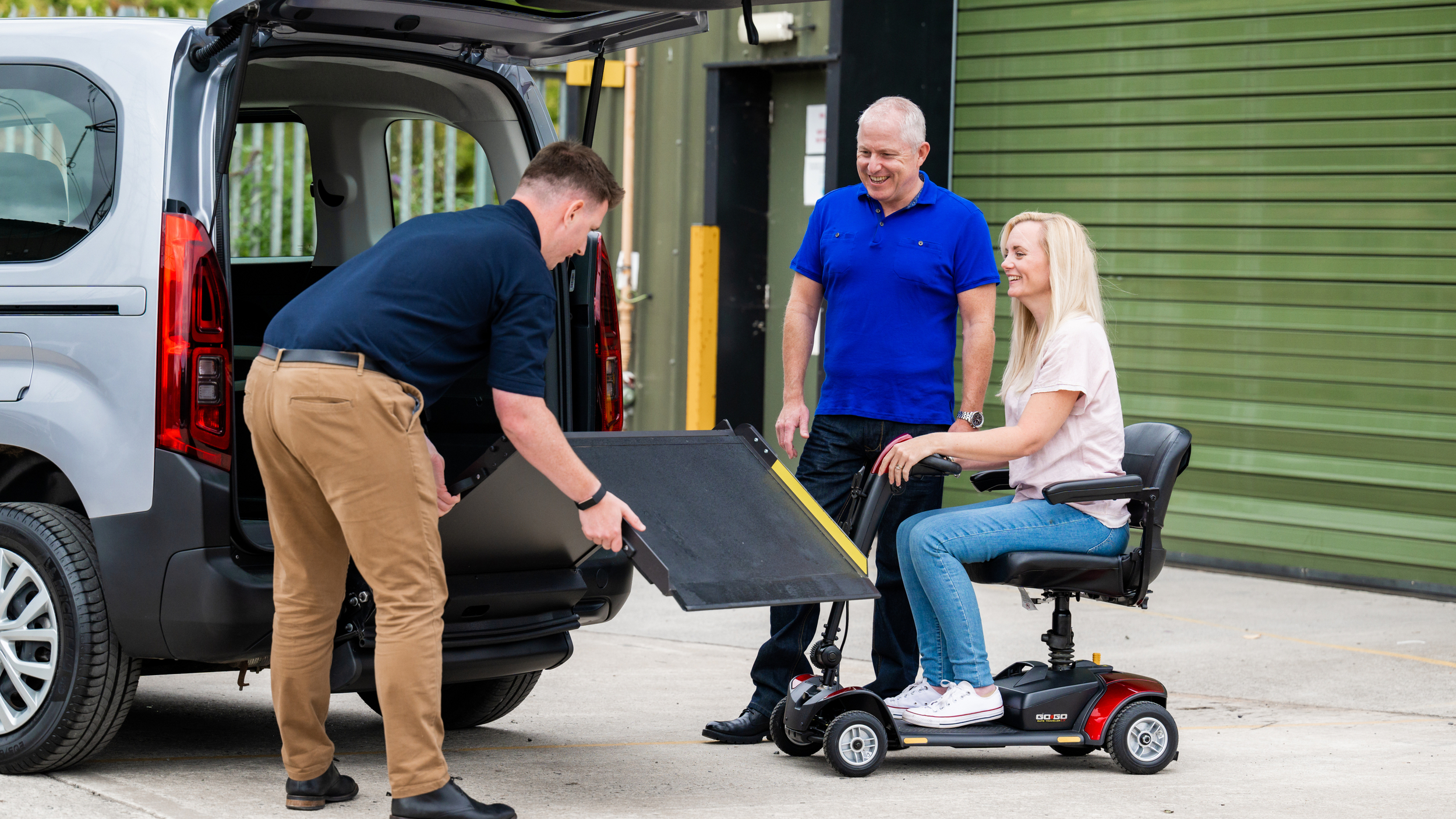 A Wheelchair Accessible Vehicle (WAV) expert lowers a ramp at the rear of the vehicle for a lady in a wheelchair and a man.
