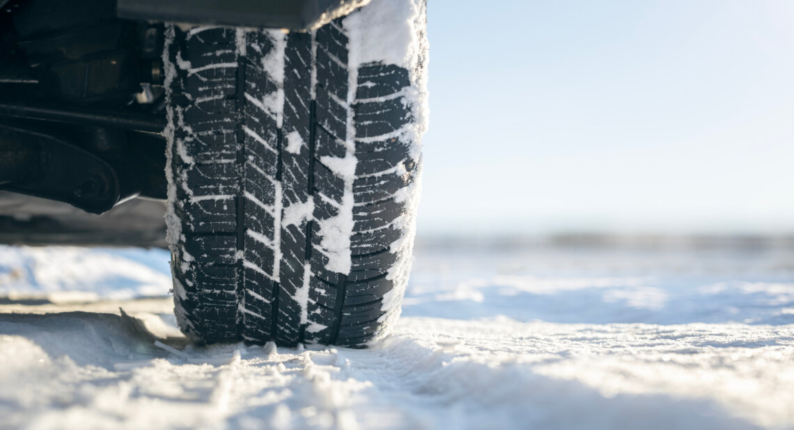 Close-up of a car tyre with snow caught in its tread, parked on a snowy road under a clear sky.