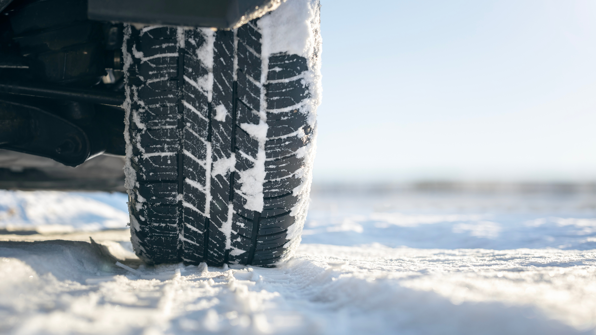 Close-up of a car tyre with snow caught in its tread, parked on a snowy road under a clear sky.