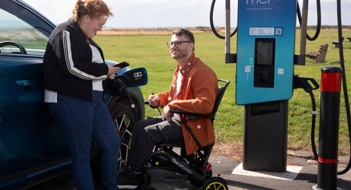 A woman leaning against a blue electric vehicle checks her phone while a man using a powered wheelchair sits beside her at a public charging point in an open grassy area. The charging cable is connected to the car and both people appear relaxed and engaged.