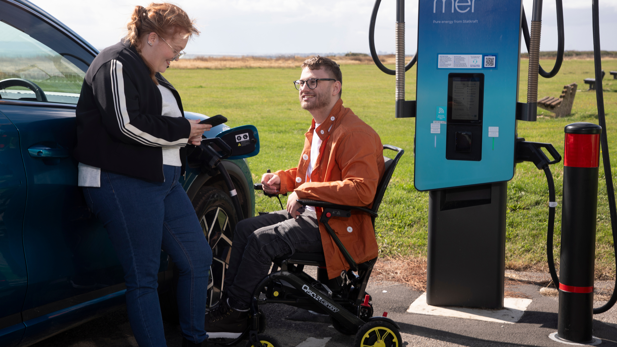 A woman leaning against a blue electric vehicle checks her phone while a man using a powered wheelchair sits beside her at a public charging point in an open grassy area. The charging cable is connected to the car and both people appear relaxed and engaged.