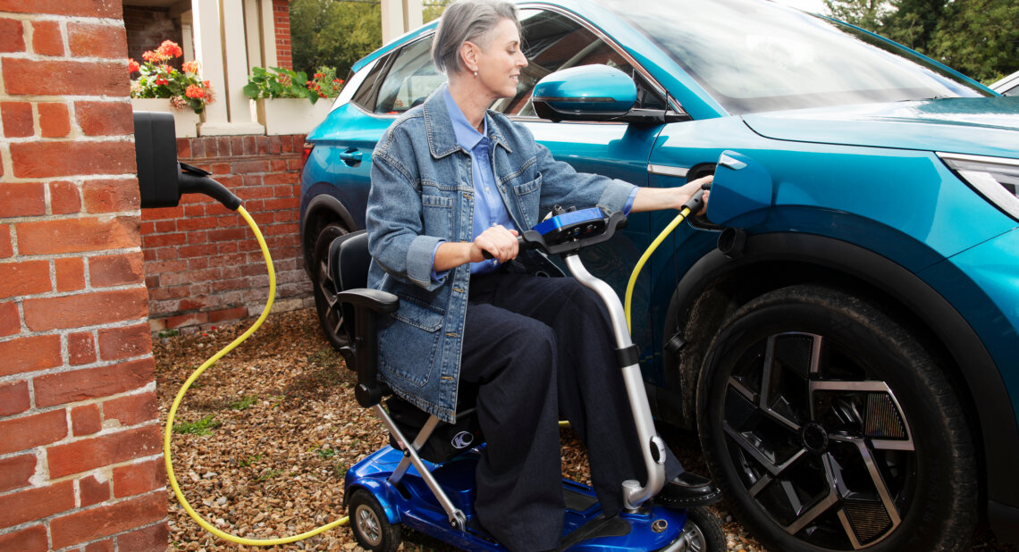 A woman using a blue mobility scooter plugs a yellow charging cable into a blue electric vehicle parked beside a red brick house. She smiles as she connects the car to the home charging point.