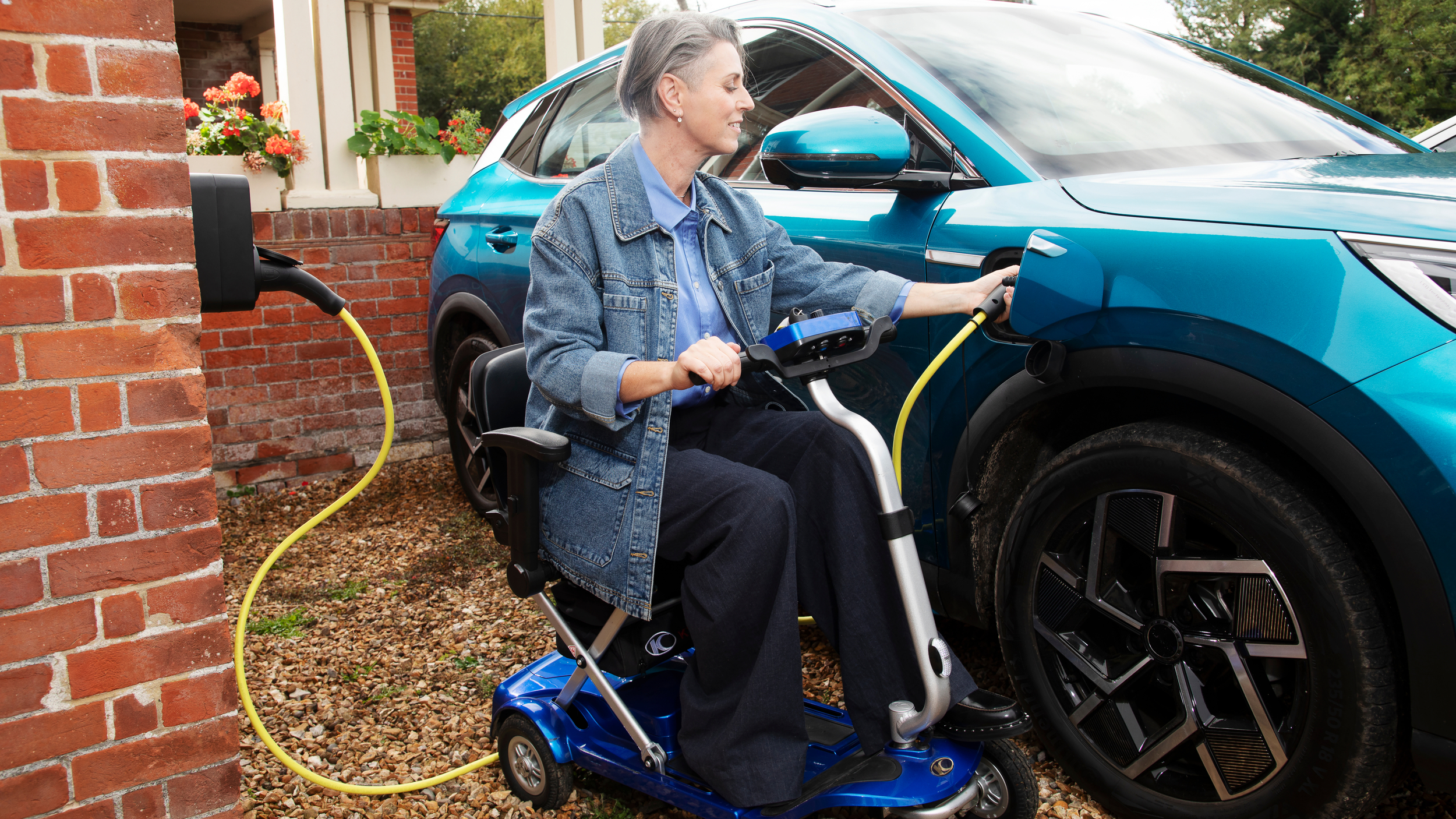 A woman using a blue mobility scooter plugs a yellow charging cable into a blue electric vehicle parked beside a red brick house. She smiles as she connects the car to the home charging point.