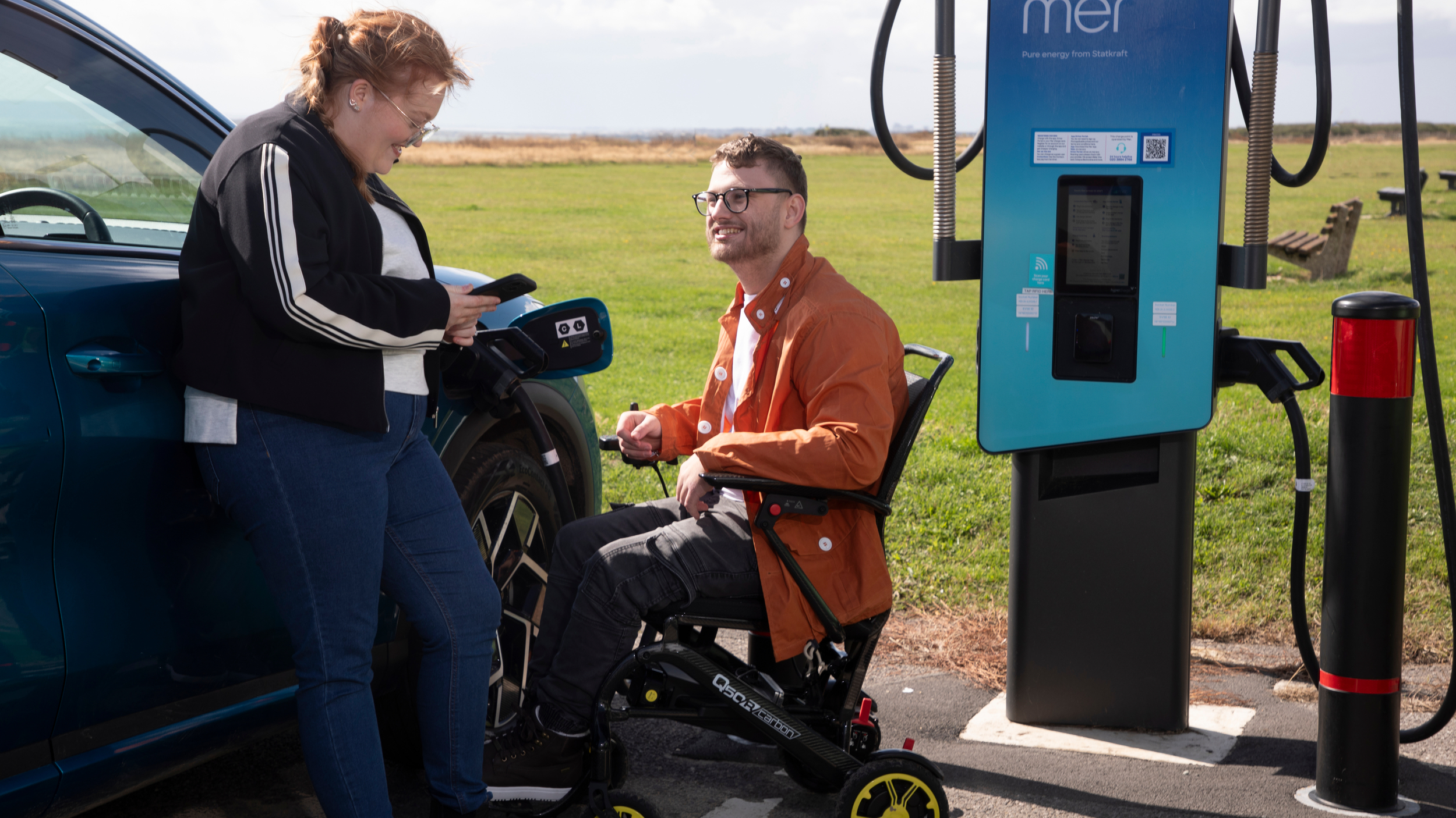 A woman stands beside a blue electric car that is plugged into a public charging point, looking at her phone and smiling. Next to her, a man in a powered wheelchair smiles up at her while sitting beside the car. The charging unit is tall and blue with cables hanging on either side. They are outdoors on a sunny day with grass and benches in the background.