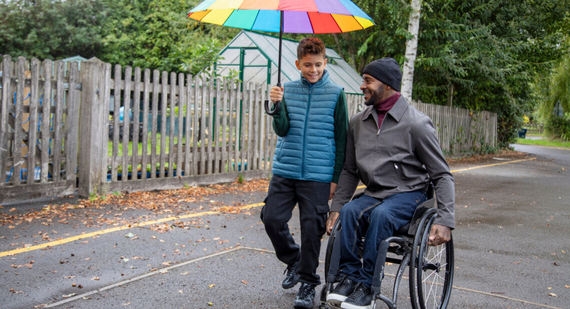 A man using a wheelchair smiles as he moves along a wet pavement beside a young boy who is holding a colourful rainbow umbrella over them. They appear to be chatting while walking through a leafy outdoor area with wooden fencing and a greenhouse in the background.