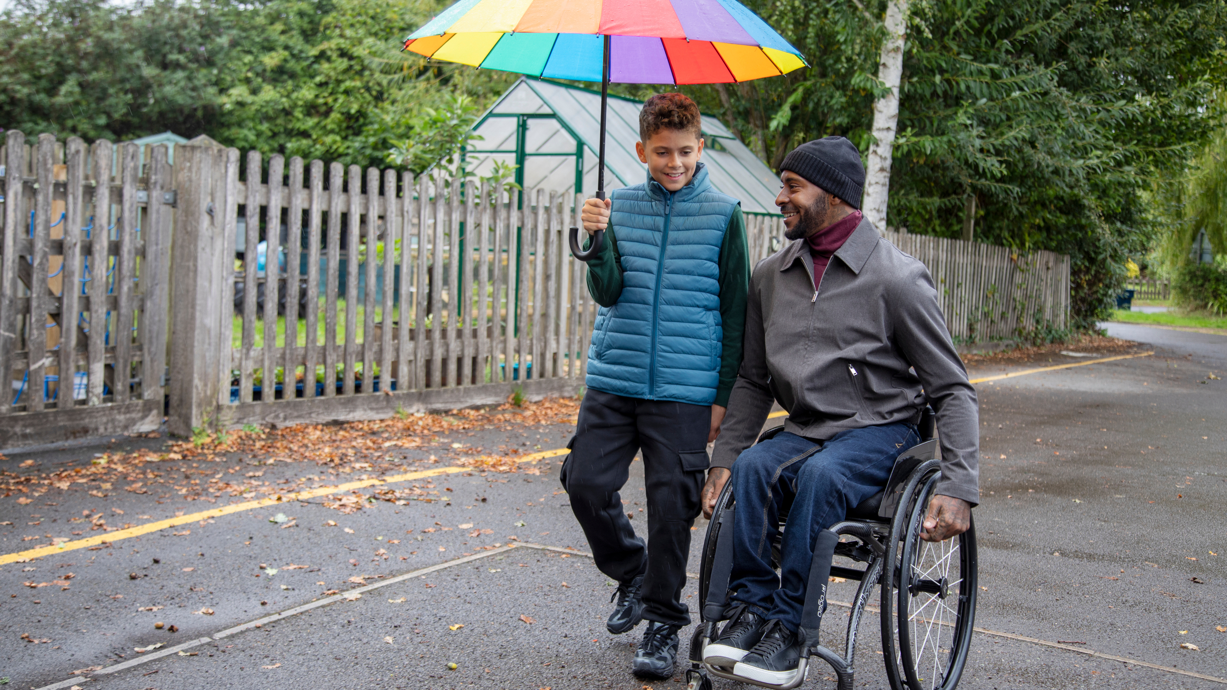 A man using a wheelchair smiles as he moves along a wet pavement beside a young boy who is holding a colourful rainbow umbrella over them. They appear to be chatting while walking through a leafy outdoor area with wooden fencing and a greenhouse in the background.