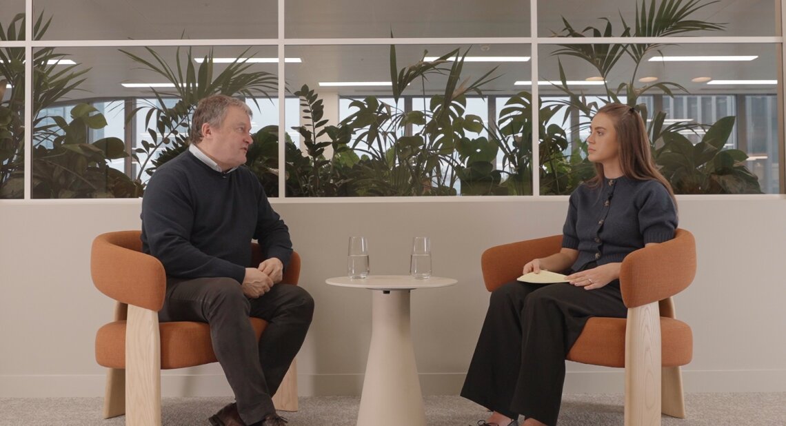 Andrew Miller, CEO of Motability Operations, sits on the left answering frequently asked customer questions about the 2025 UK Autumn Budget, while Morgan from the Customer Services team sits on the right. They talk across a small round table with two glasses of water, with large indoor plants visible behind a glass partition.
