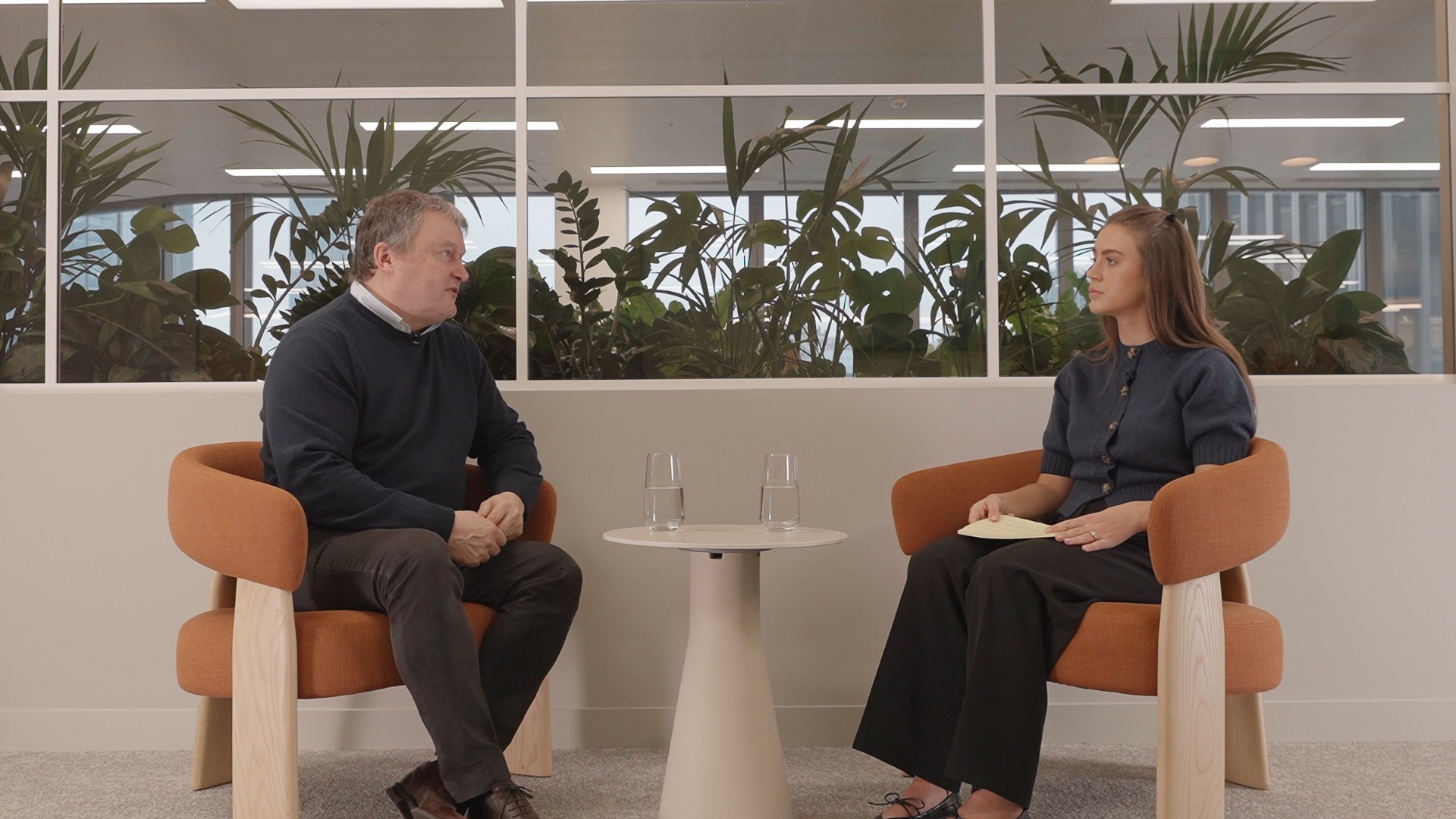 Andrew Miller, CEO of Motability Operations, sits on the left answering frequently asked customer questions about the 2025 UK Autumn Budget, while Morgan from the Customer Services team sits on the right. They talk across a small round table with two glasses of water, with large indoor plants visible behind a glass partition.