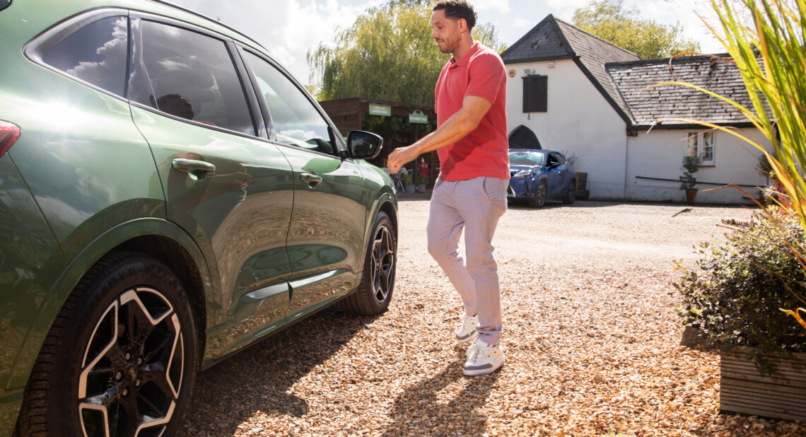 A man wearing a coral polo shirt and light trousers walks toward a green SUV parked on a gravel drive, with a white cottage and trees in the background on a bright sunny day.