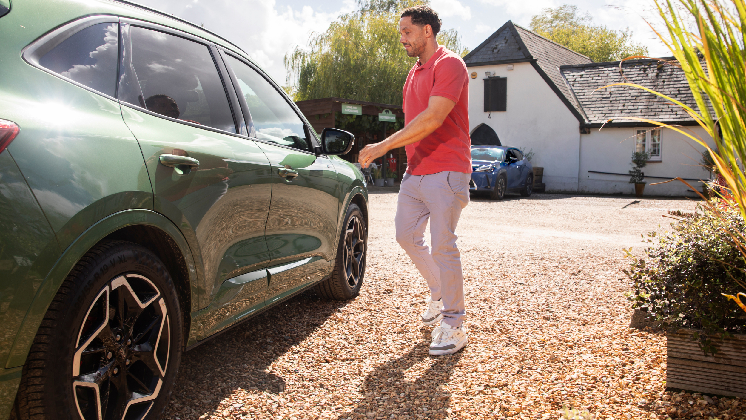 A man wearing a coral polo shirt and light trousers walks toward a green SUV parked on a gravel drive, with a white cottage and trees in the background on a bright sunny day.