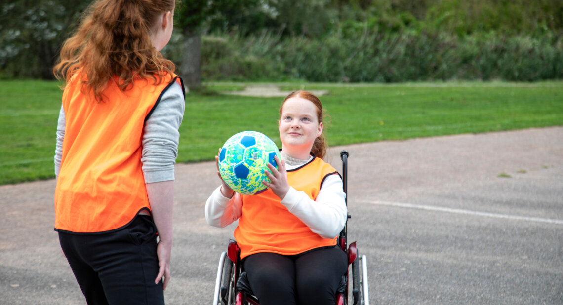 A teenage girl in a wheelchair holds a bright blue football while looking up at another girl standing beside her. Both wear orange training bibs on an outdoor sports court with grass and trees in the background.
