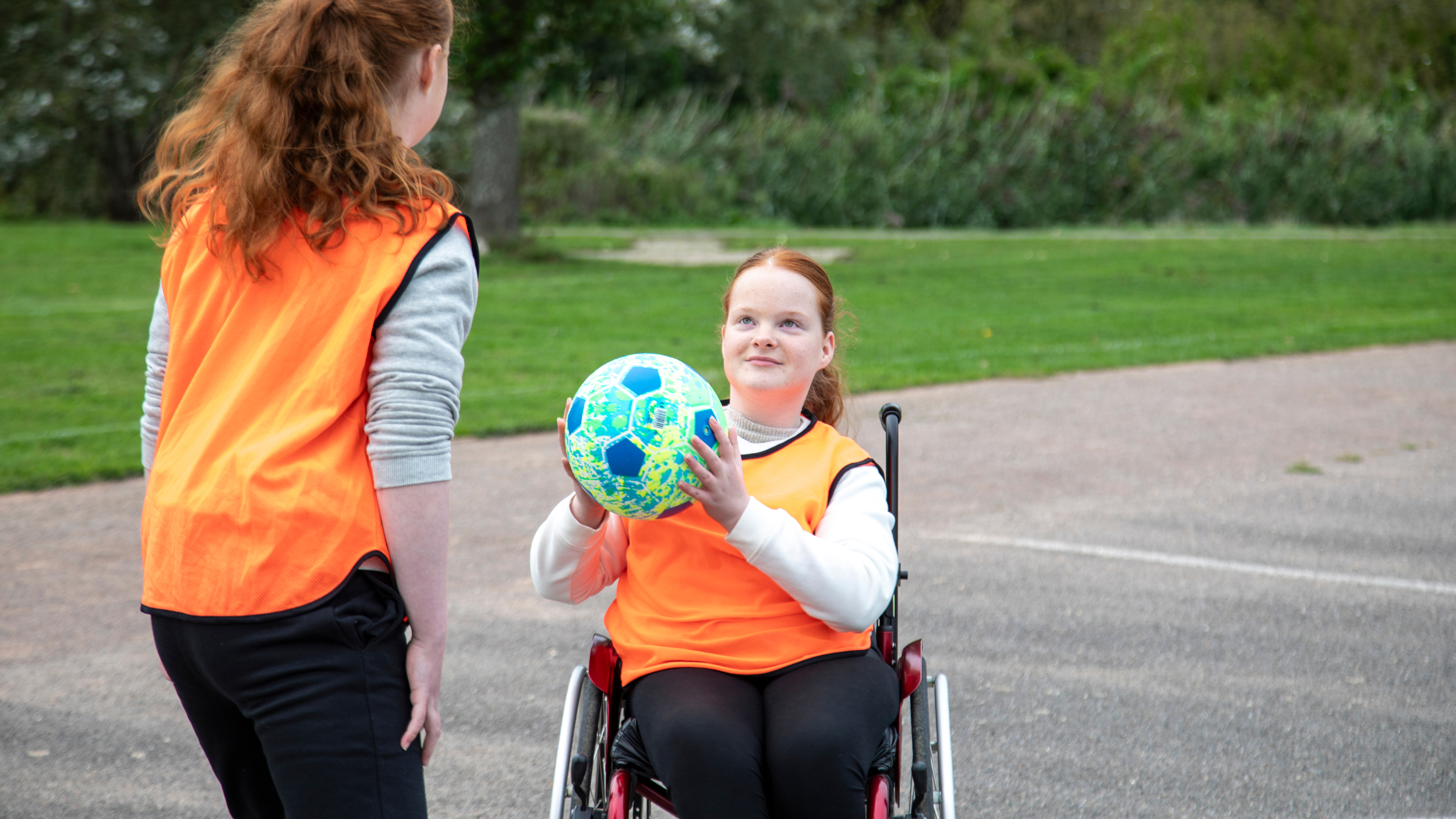 A teenage girl in a wheelchair holds a bright blue football while looking up at another girl standing beside her. Both wear orange training bibs on an outdoor sports court with grass and trees in the background.