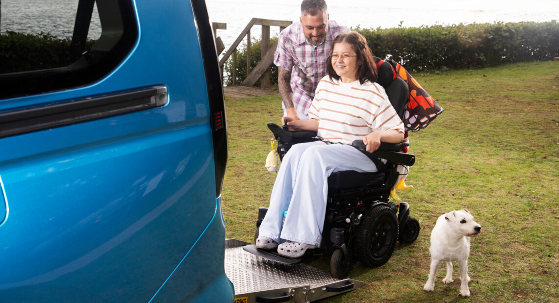 A young woman in a powered wheelchair smiles as she is guided up a lowered ramp into a blue wheelchair accessible vehicle beside a lakeside. A man stands behind her offering support and a small white dog waits nearby.