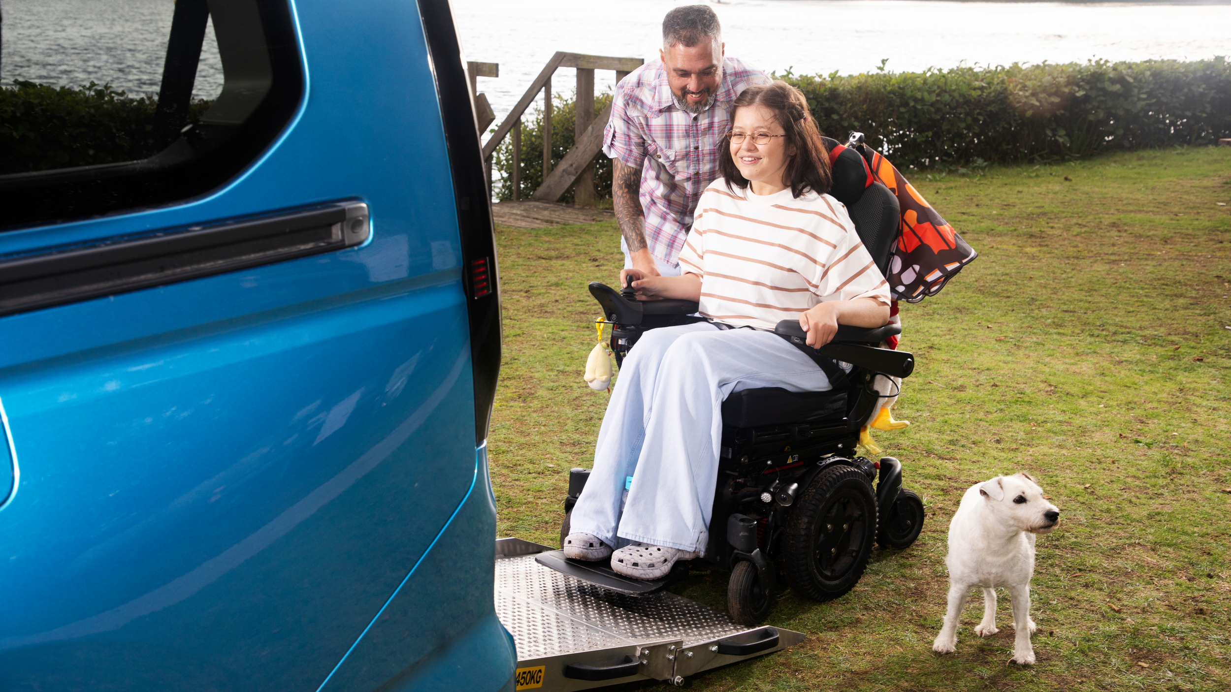 A young woman in a powered wheelchair smiles as she is guided up a lowered ramp into a blue wheelchair accessible vehicle beside a lakeside. A man stands behind her offering support and a small white dog waits nearby.