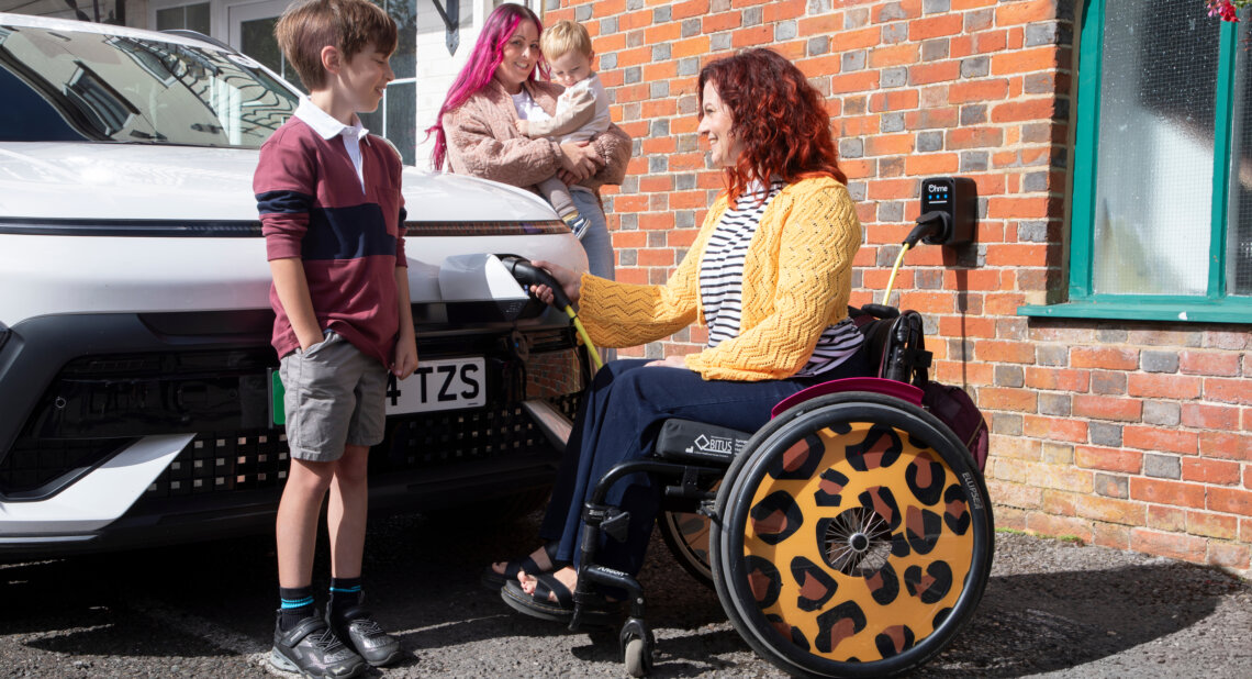 A woman in a wheelchair with leopard print wheel covers plugs an electric car into a wall-mounted charger outside a brick house. She smiles at a young boy standing beside the car, while another woman holding a toddler looks on from behind.