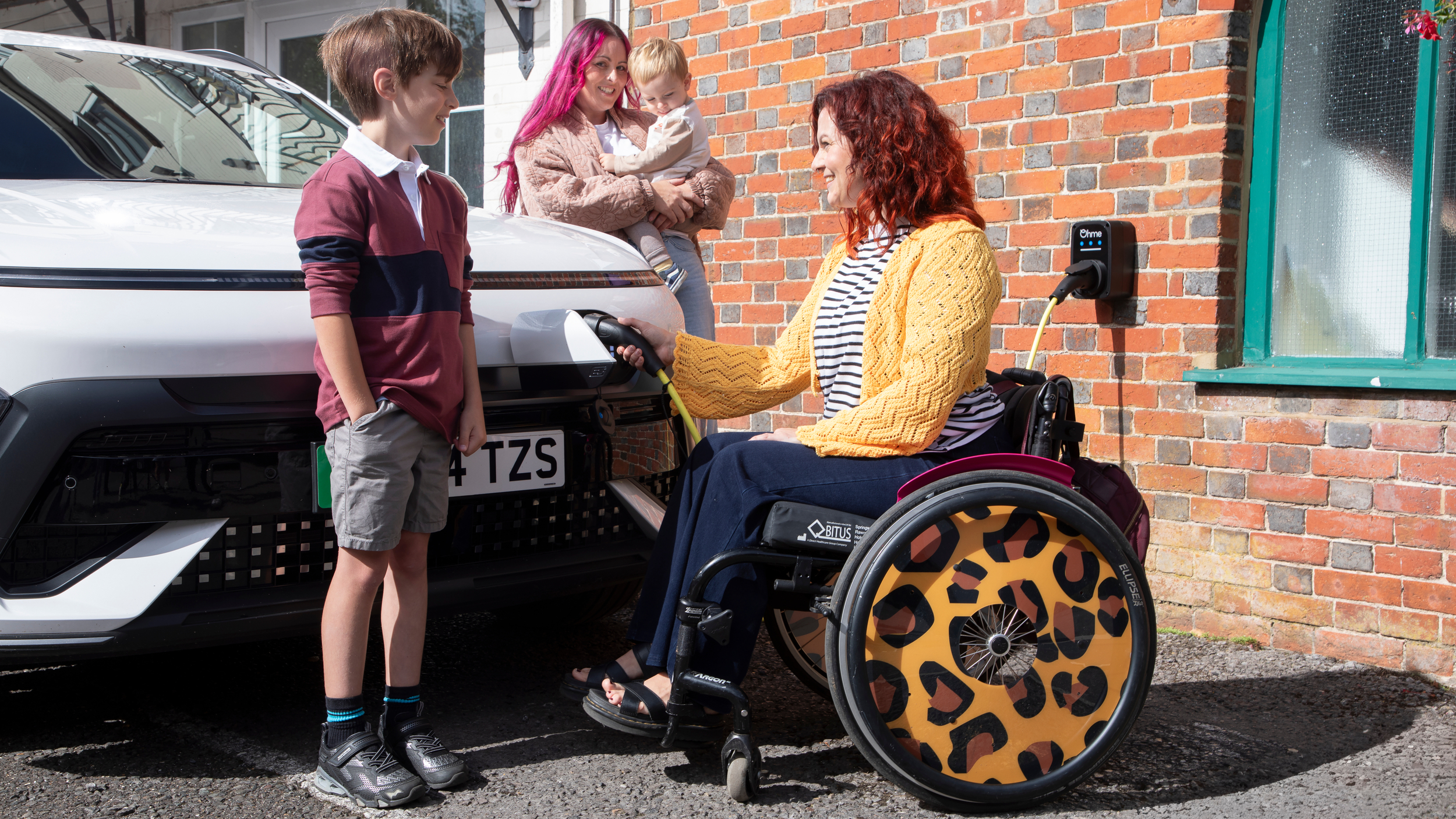 A woman in a wheelchair with leopard print wheel covers plugs an electric car into a wall-mounted charger outside a brick house. She smiles at a young boy standing beside the car, while another woman holding a toddler looks on from behind.
