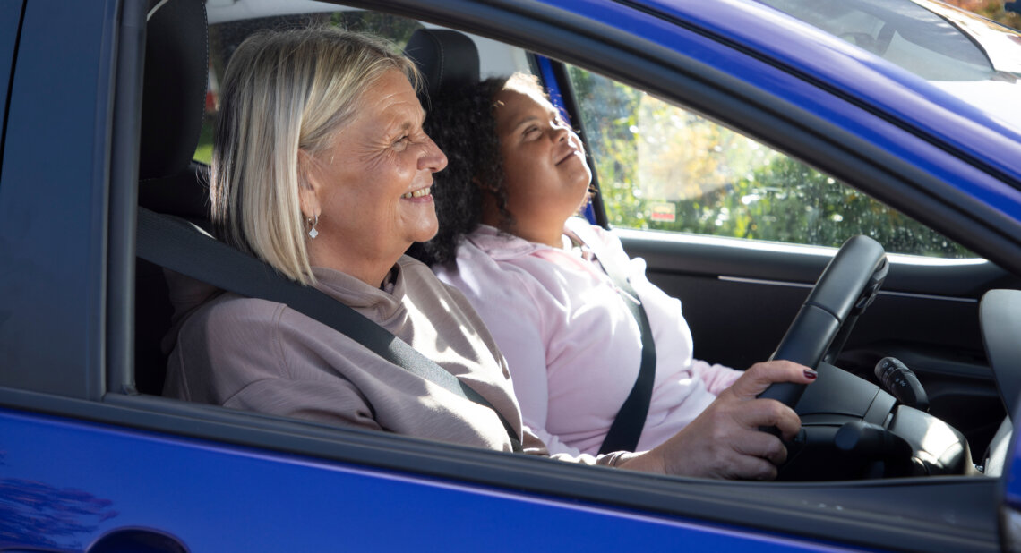 Two women sit in the front of a blue car on a sunny day. The driver smiles while holding the steering wheel and the passenger looks upward with a relaxed expression. Sunlight shines through the windows and greenery is visible outside.