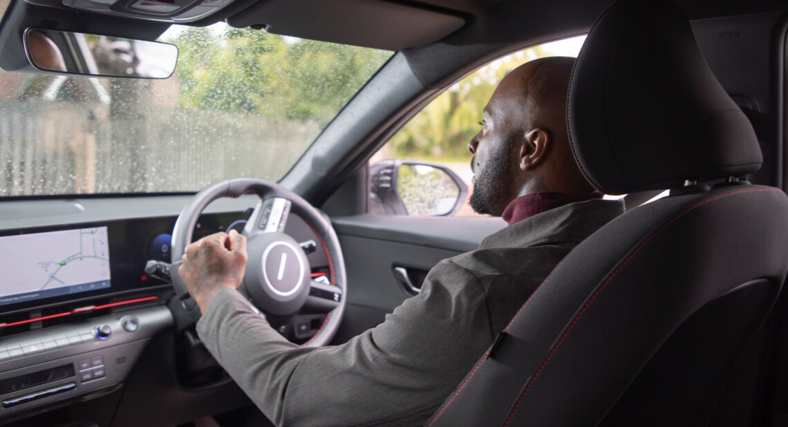 A man driving a Motability Scheme car, using a hand control, viewed from the back seat, with rain visible on the side window and a navigation screen displayed on the dashboard.