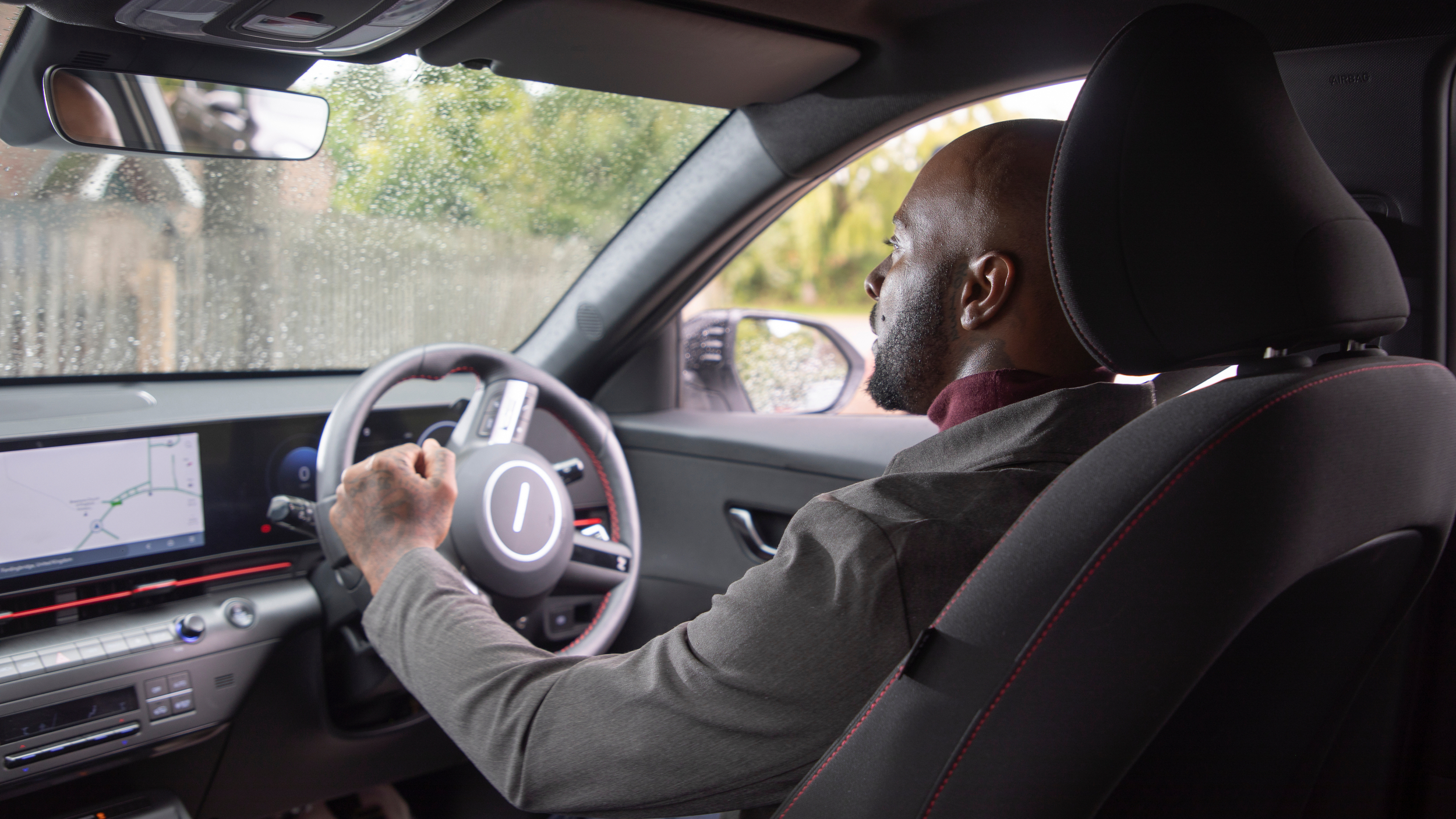 A man driving a Motability Scheme car, using a hand control, viewed from the back seat, with rain visible on the side window and a navigation screen displayed on the dashboard.