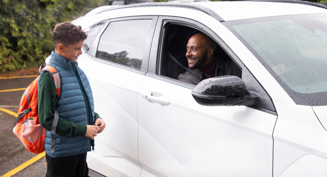 A man sits inside a car, looking out through the driver's windscreen at a child who is stood outside looking back at him.