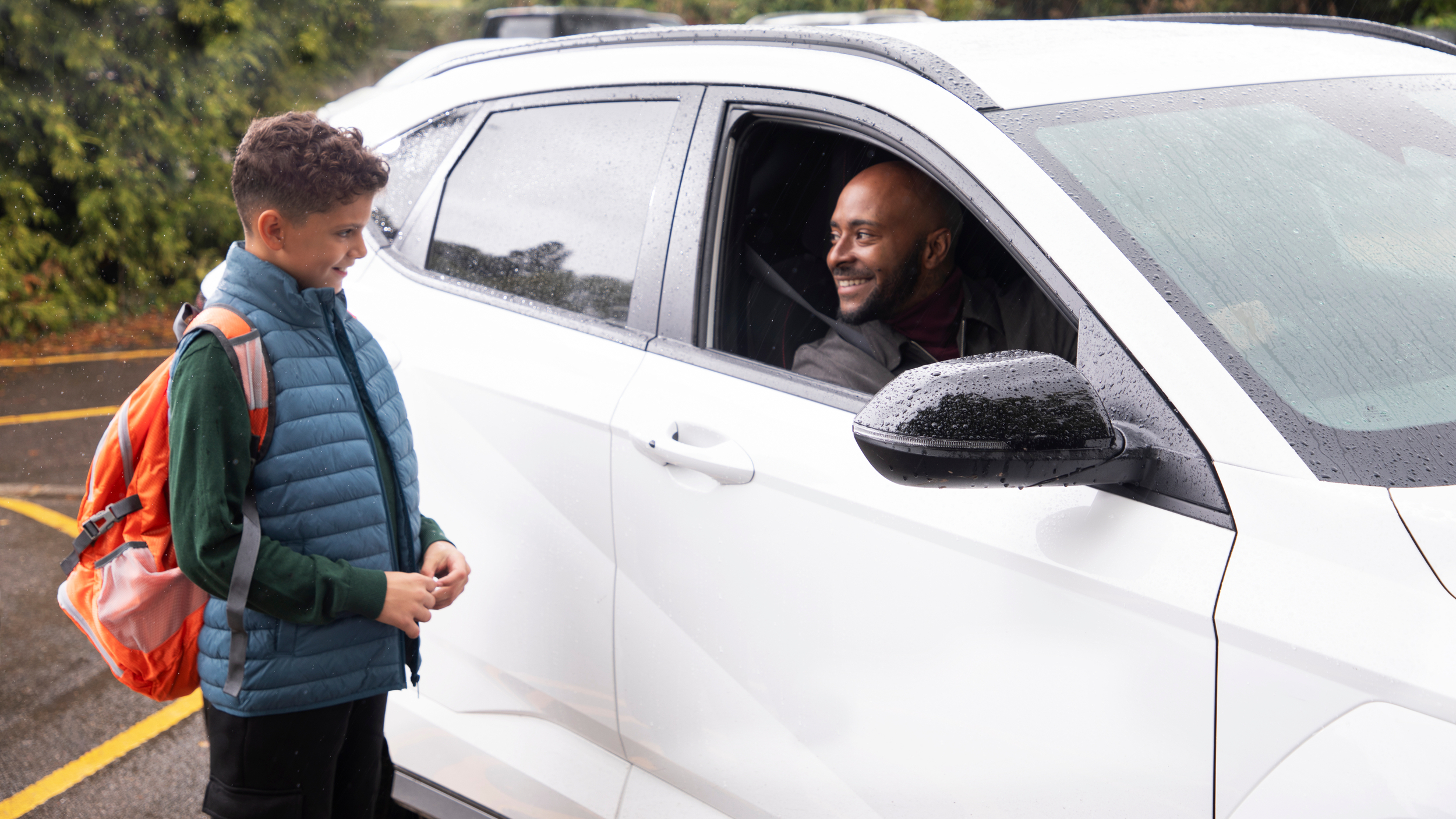 A man sits inside a car, looking out through the driver's windscreen at a child who is stood outside looking back at him.