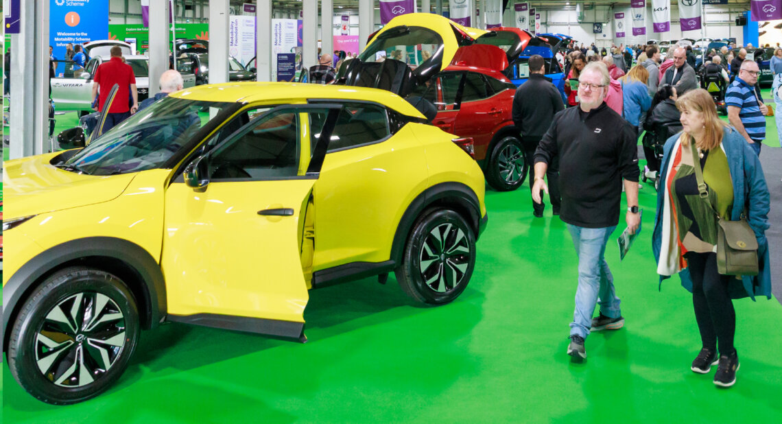 Visitors walk through a busy Motability Scheme exhibition hall, passing a bright yellow car with its door and boot open for viewing.